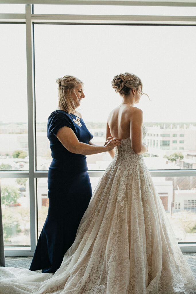 The mother of the bride assists her daughter with the buttons on the back of her lace wedding gown before her elopement in Texas