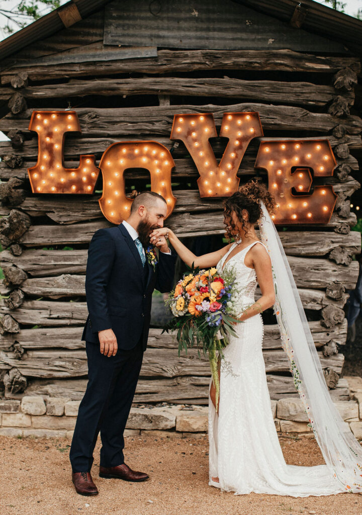 A groom kisses his bride's hand in front of a lit sign at their Austin TX wedding