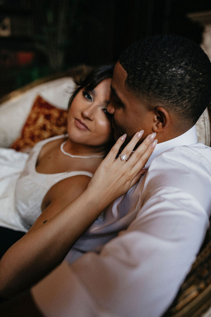An image showcases a diamond engagement ring as a couple embraces inside Chateau Nouvelle for their houston engagement photography session 