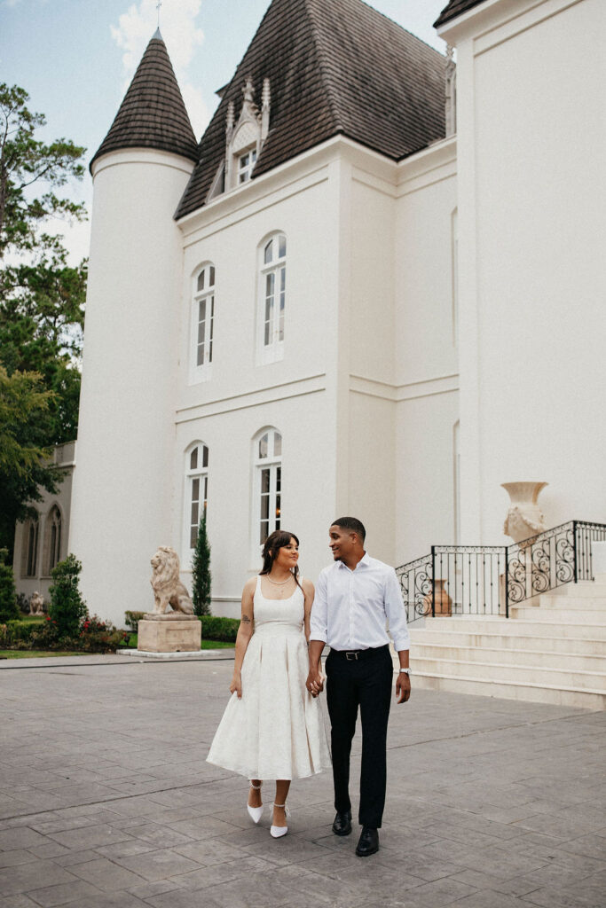A young couple walks hand-in-hand at the front exterior of Chateau Nouvelle, a French-inspired venue, for their houston engagement photography session