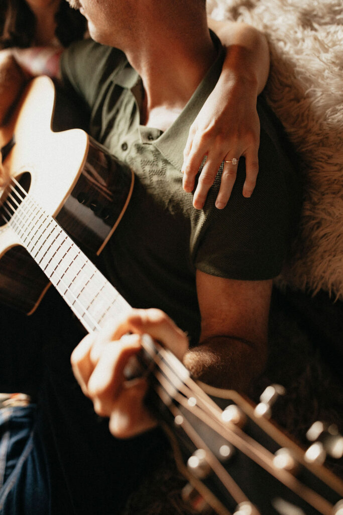 A Houston engagement photographer narrows in on a diamond engagement ring as a woman's arm is draped over her fiancé's shoulder while he plays guitar
