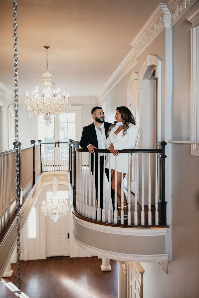 A Houston couple smile next to a chandelier inside Sandlewood Manor for their engagement photos in Houston TX 