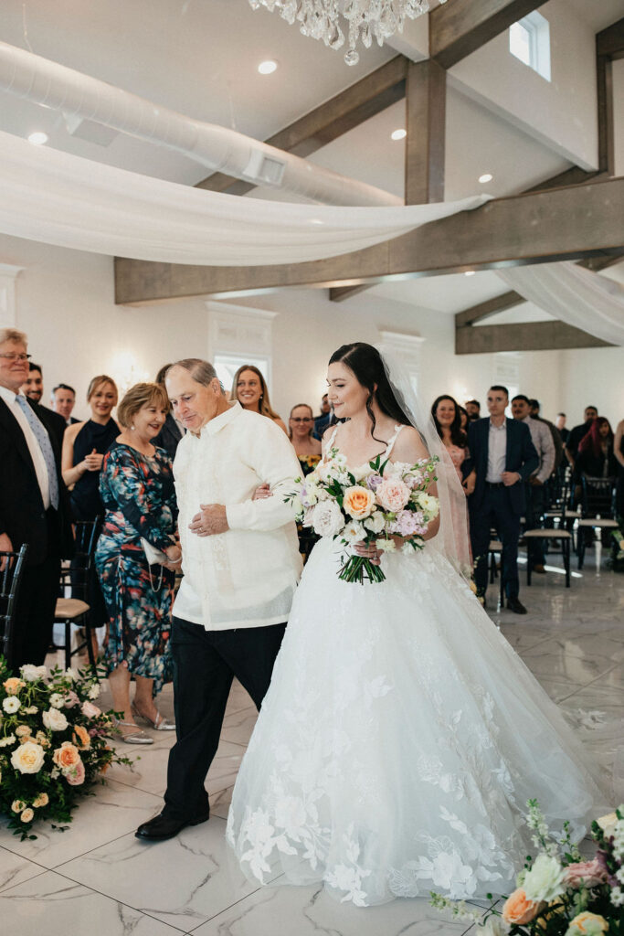 A bride is escorted down the aisle wearing a lace wedding gown at her spring wedding at Sandlewood Manor.