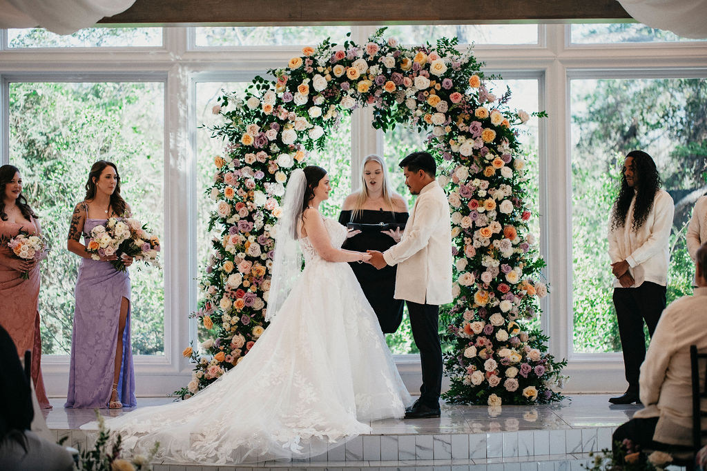 A bride & groom hold hands as they exchange vows beneath a spring floral arch at their Sandlewood Manor wedding. 