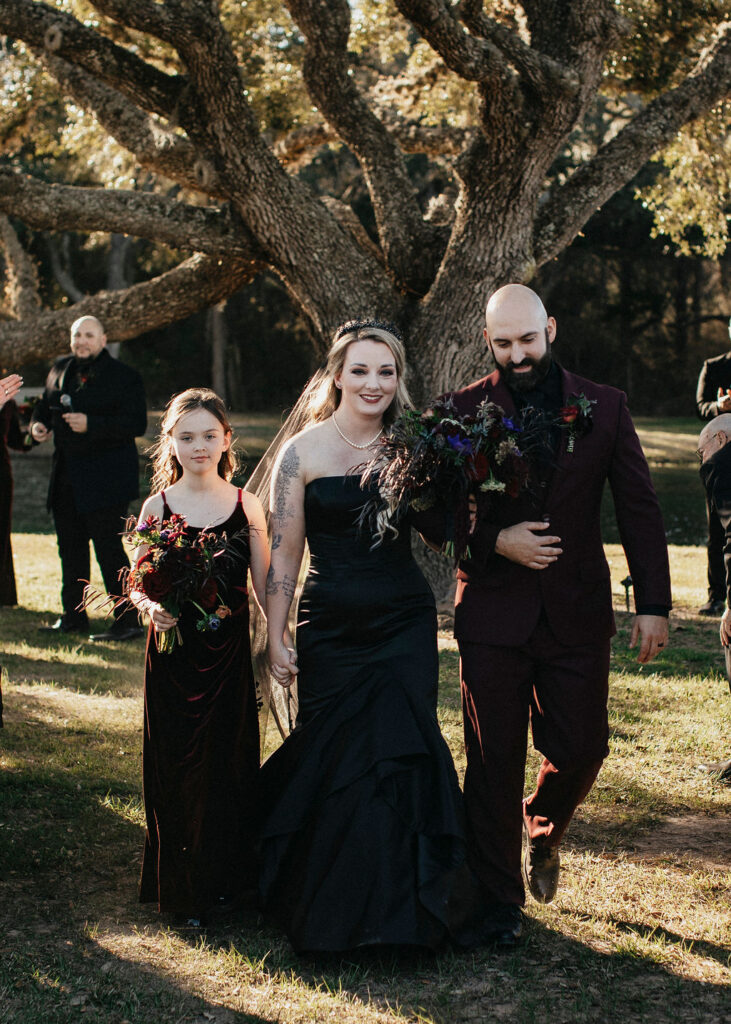 A bride wearing a black wedding dress is escorted down the aisle with her husband and daughter at their m3 ranch wedding.