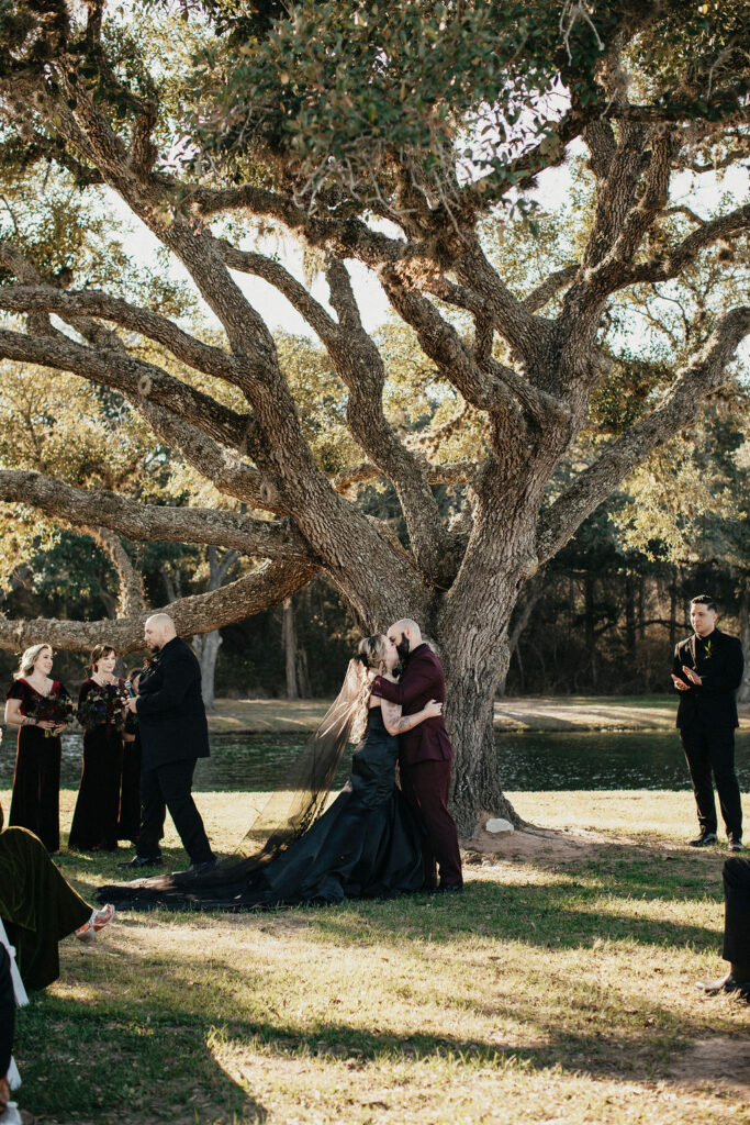 A bride and groom share a kiss beneath a tree at their outdoor ceremony at a Texas ranch wedding venue.